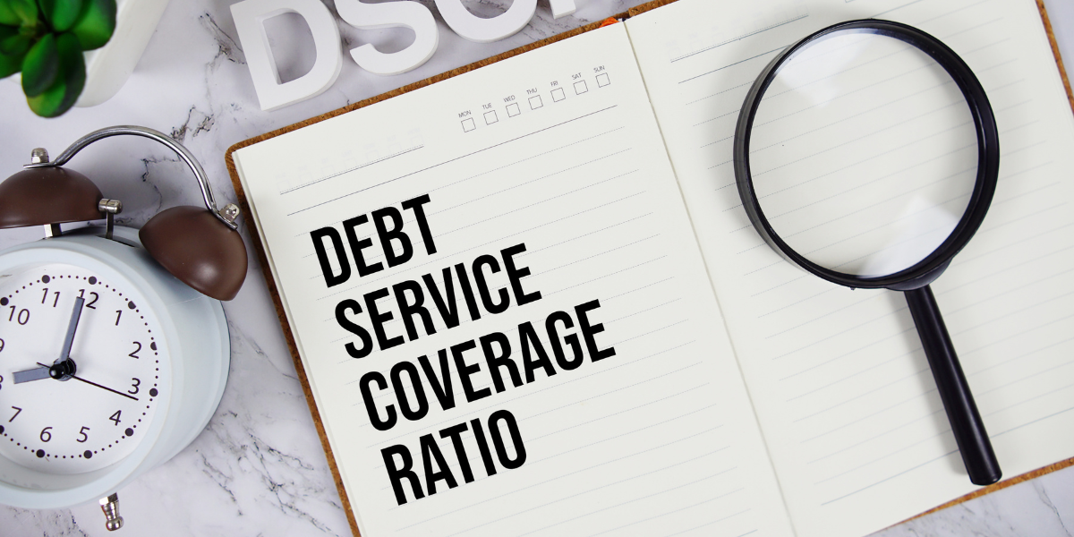 text in an open notebook defining Debt Service Coverage Ratio (DSCR), featuring a magnifying glass and an alarm clock on a marble desk.